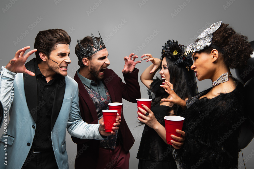 multiethnic friends in halloween costumes frightening each other isolated on grey