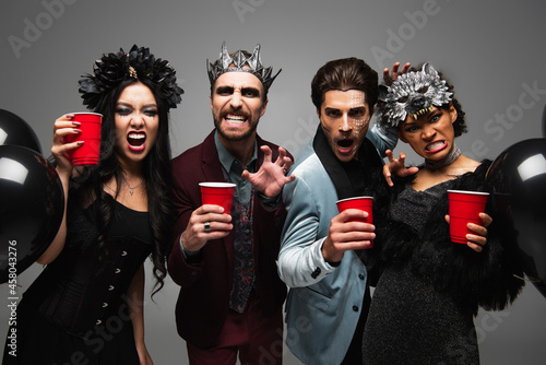 spooky multiethnic friends with plastic cups growling and showing scary gesture isolated on grey