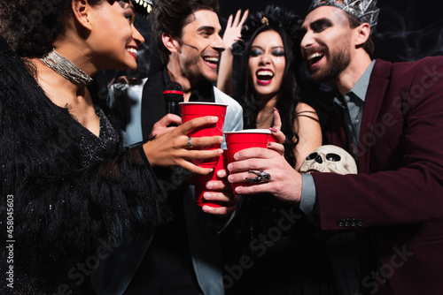 cheerful multiethnic friends toasting with plastic cups while singing karaoke on black