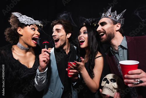 Canvas Print joyful multiethnic friends in spooky costumes singing karaoke on halloween party