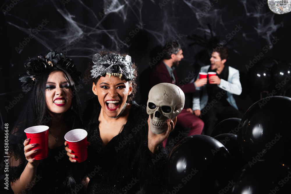 interracial women in spooky halloween costumes grinning and growling near blurred friends talking on black