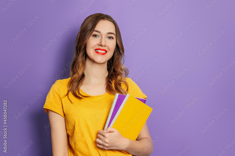 Caucasian student girl holding a book, notebook smiling, looking at the camera isolated on lilac background in studio Caucasian student girl holding a book, notebook smiling, looking at the camera isolated on lilac background in studio