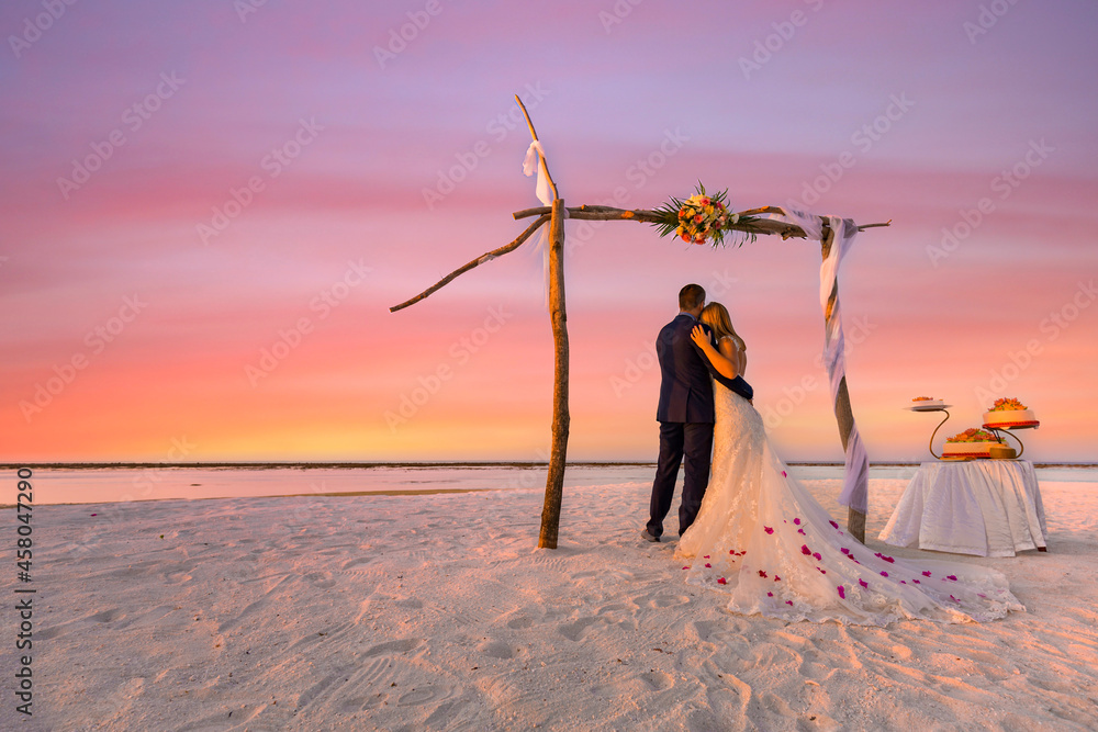 Bride and groom, newlyweds, honeymoon on the beach sunset sun under wedding arch with cake. Lovers or newlywed married young couple by the sea, wedding ceremony on a tropical beach, ocean background - obrazy, fototapety, plakaty 