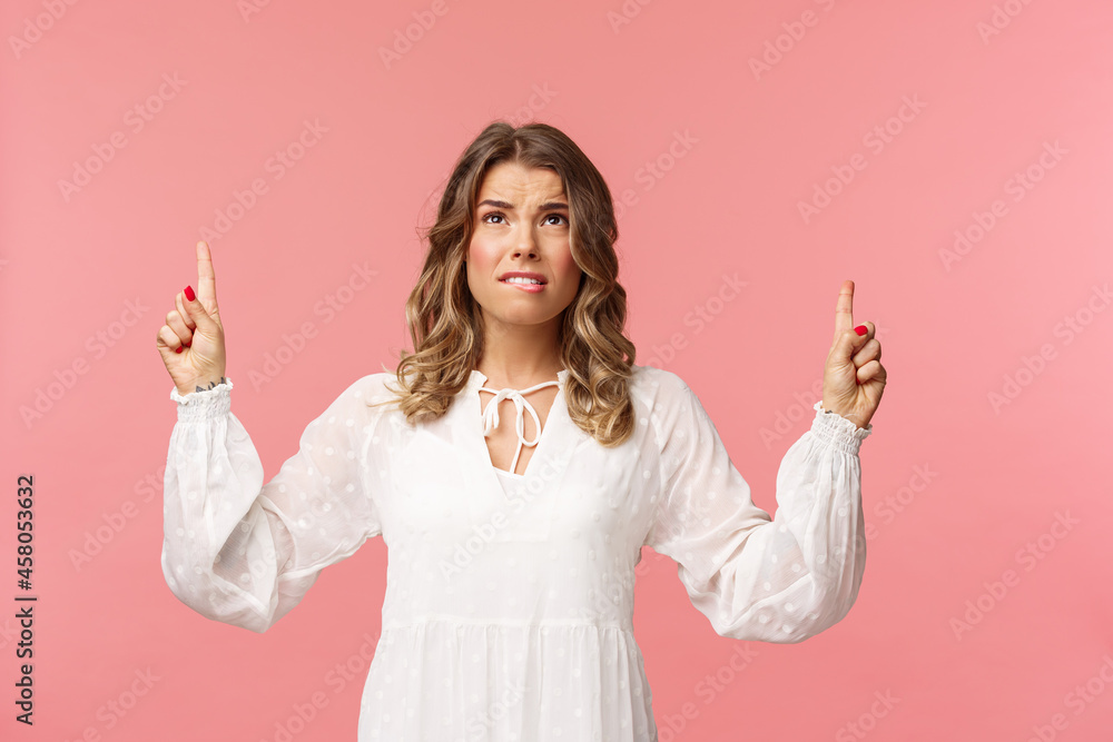 Skeptical and embarrassed blond caucasian woman in white cute dress, grimacing, cringe from seeing something disgusting, frowning doubtful and pointing fingers up strange thing, pink background - obrazy, fototapety, plakaty 
