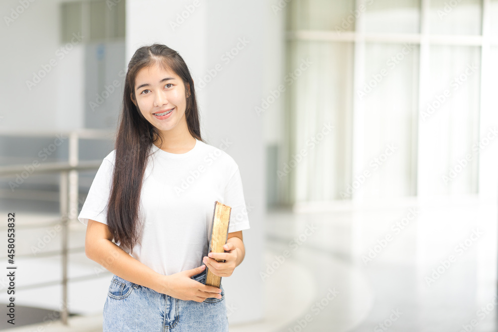 Front view portrait of a young cheerful Asian adolescent woman college student in relaxed casual back to school carry books stands in campus building