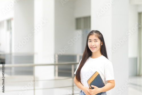 Front view portrait of a young cheerful Asian adolescent woman college student in relaxed casual back to school carry books stands in campus building