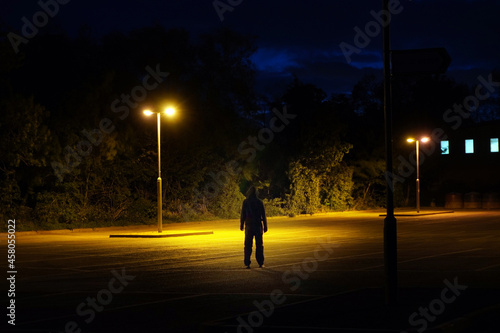 A hooded figure standing below street lights in a car park. On a summers night. © Dave