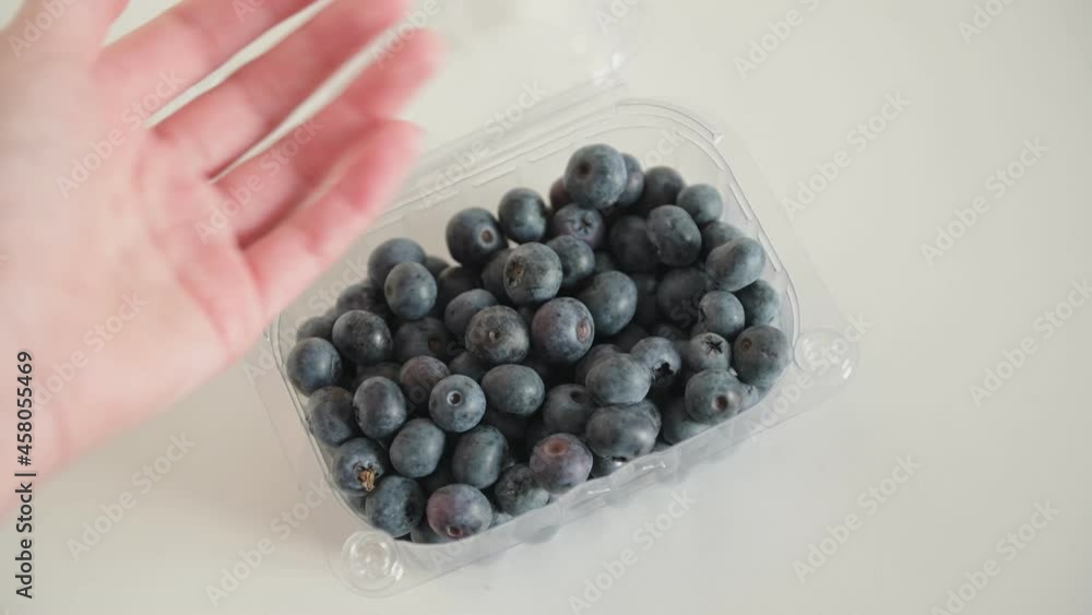 Woman hand with long nails takes blueberries from plastic container standing on white surface and puts berries back closeup Woman hand with long nails takes blueberries from plastic container standing on white surface and puts berries back closeup