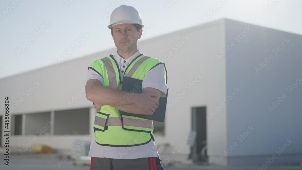 Zoom in to serious confident professional Caucasian man in uniform and hard hat with crossed hands looking at camera posing outdoors on sunny day at warehouse. Expert storage manager in sunshine Zoom in to serious confident professional Caucasian man in uniform and hard hat with crossed hands looking at camera posing outdoors on sunny day at warehouse. Expert storage manager in sunshine