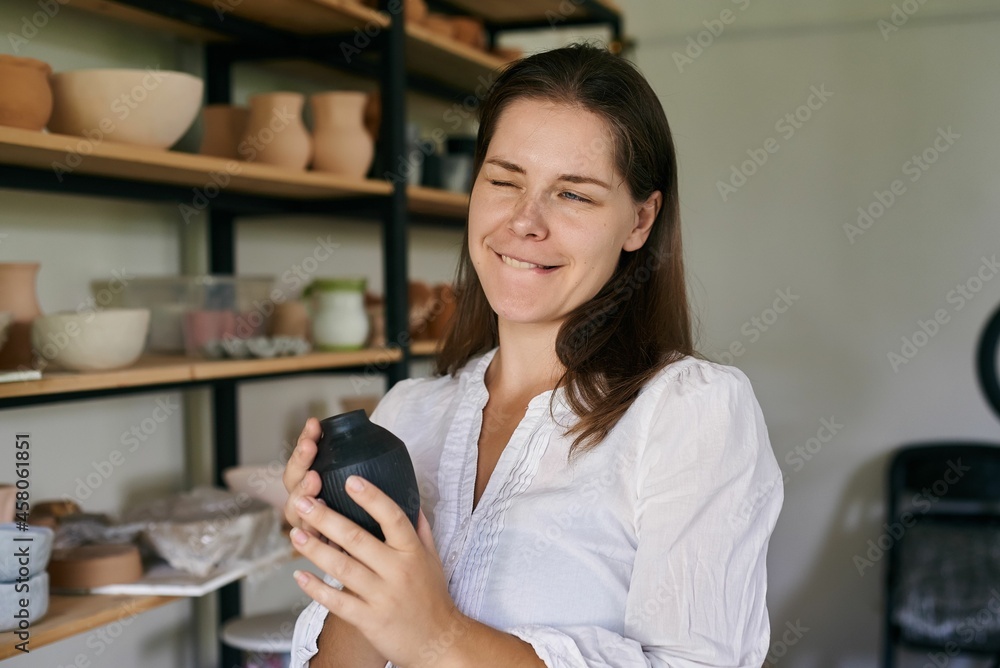 woman artisan ceramist stands against rack handmade clay utensils, holds small black vase