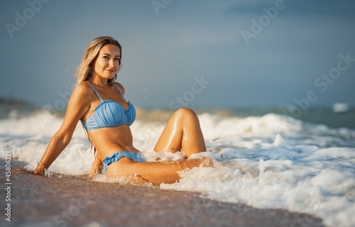 A girl with blond hair in a delicate swimsuit sits by the sea, enjoying the splash of waves