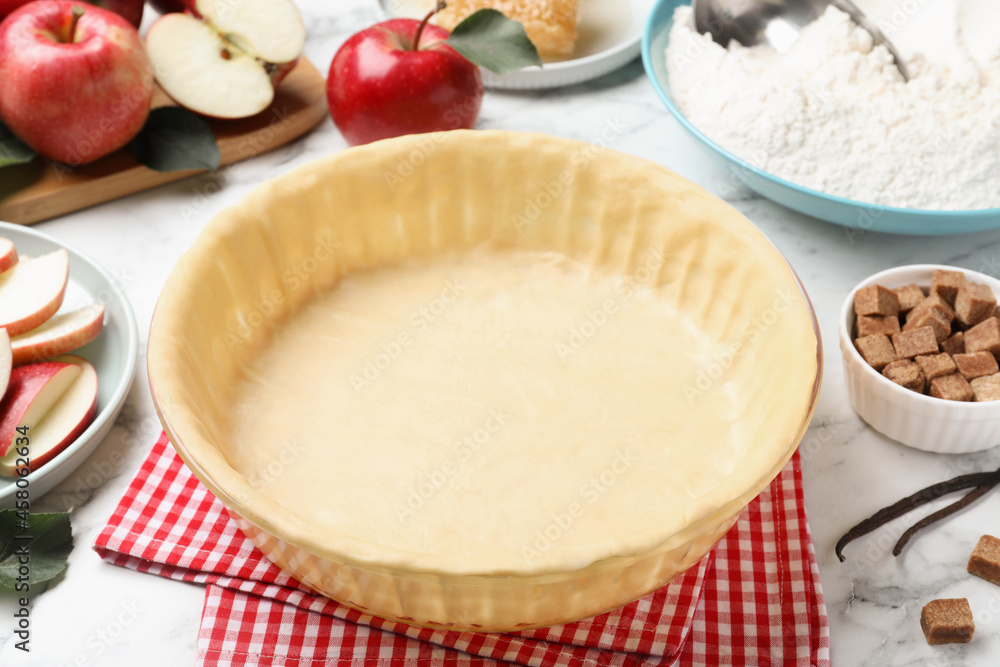 Baking dish with raw dough for apple pie and ingredients on white marble table Baking dish with raw dough for apple pie and ingredients on white marble table