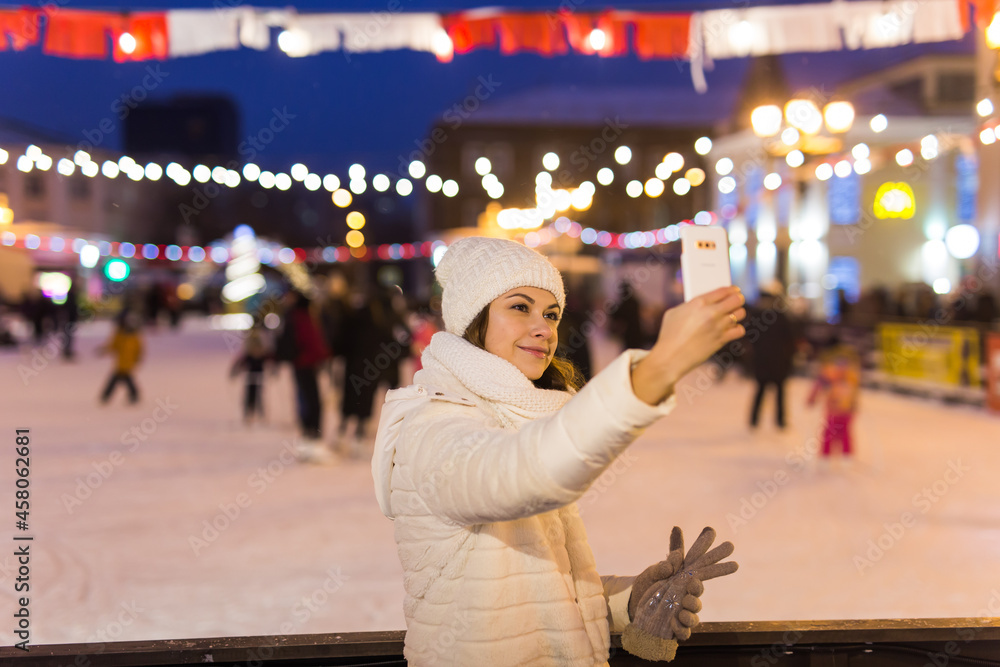 Obraz premium Christmas, winter, technology and leisure concept - happy young woman taking picture with smartphone on ice skating rink outdoors.