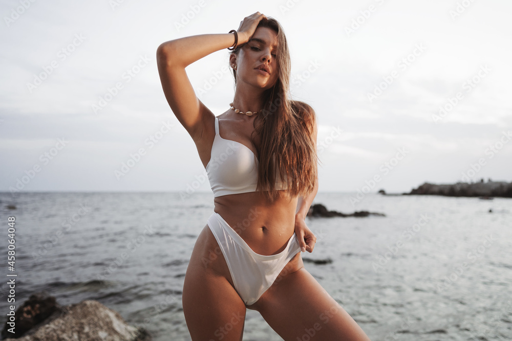 Fototapeta premium Sexy beautiful woman in white swimwear at sea coast on sunset
