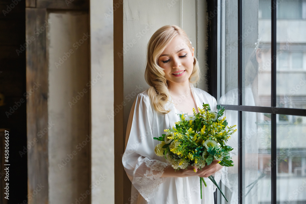 Wedding morning of the bride. Exquisite blonde in a delicate peignoir standing near the window with a bouquet of flowers in her hands Wedding morning of the bride. Exquisite blonde in a delicate peignoir standing near the window with a bouquet of flowers in her hands