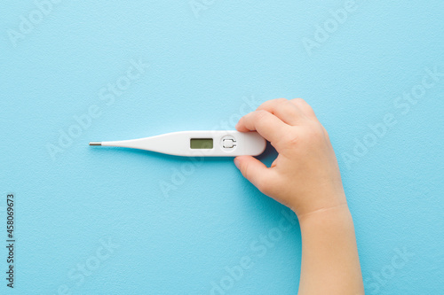 Baby hand holding white digital thermometer on light blue table background. Pastel color. Closeup. Temperature measuring of children. Healthcare concept. Top down view. photo