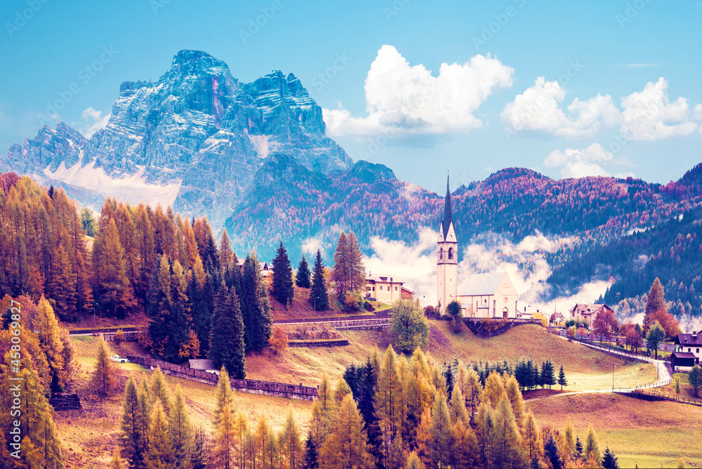 Amazing magical autumn landscape with the church and yellow larch on a background of mountains in the Dolomites in a sunny day near Codalonga, Alps, Italy.