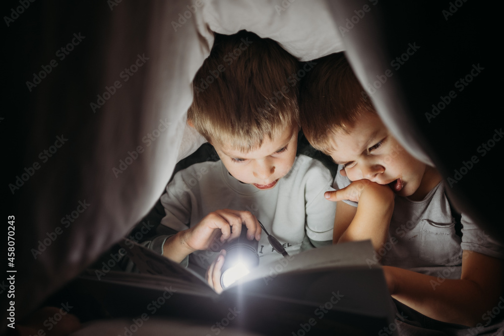 two little brothers reading a book at night hiding under a blanket two little brothers reading a book at night hiding under a blanket