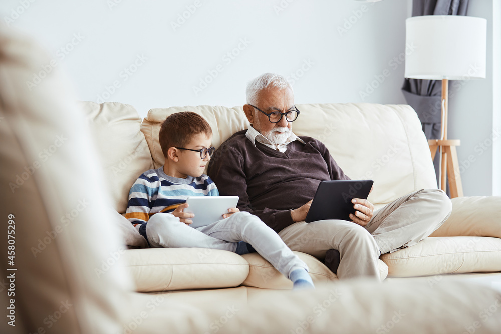 Grandfather and grandson use digital tablets while relaxing on the sofa at home. Grandfather and grandson use digital tablets while relaxing on the sofa at home.