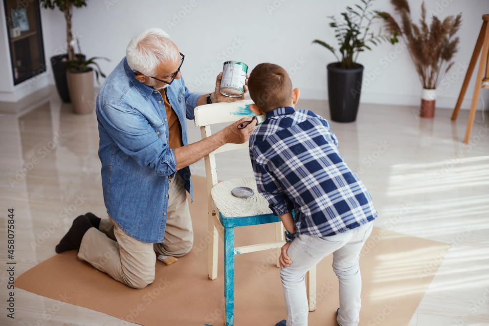 Senior man and his grandson paint chair in blue at home.