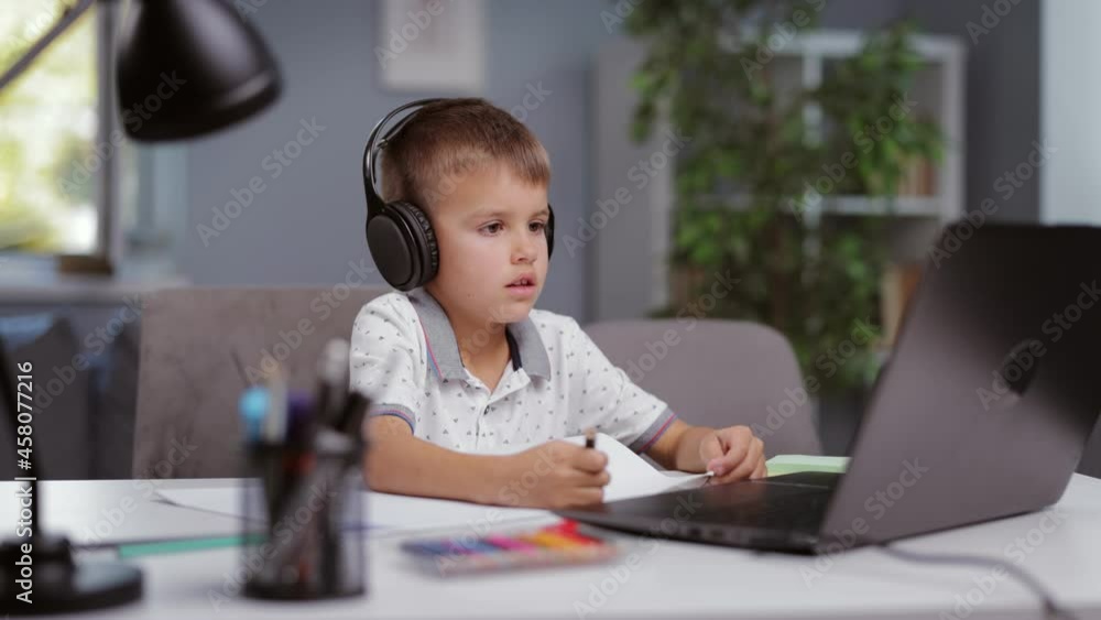 Caucasian schoolboy using wireless headphones and laptop for online lesson from home. Cute kid sitting at desk, looking at computer screen and taking notes. Distance learning concept. Caucasian schoolboy using wireless headphones and laptop for online lesson from home. Cute kid sitting at desk, looking at computer screen and taking notes. Distance learning concept.