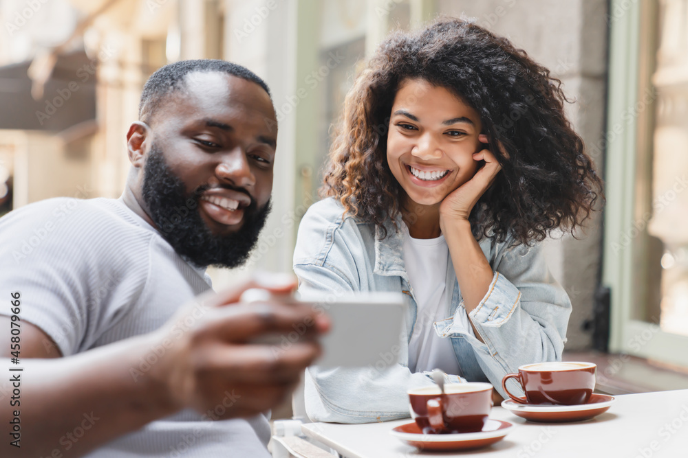 Cheerful african-american young couple friends spouses watching photos social media video on smart phone together in city cafe on romantic date