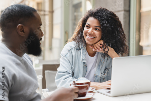Interested young african-american couple talking communicating together in cafe, drinking coffee, spending time together on romantic date using laptop for remote work.