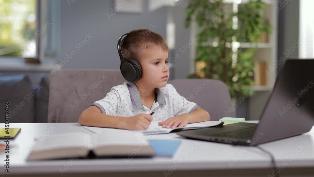 Concentrated boy sitting at desk in wireless headphones and taking notes during online lesson on modern laptop. Caucasian pupil having distance education while staying at home. Concentrated boy sitting at desk in wireless headphones and taking notes during online lesson on modern laptop. Caucasian pupil having distance education while staying at home.