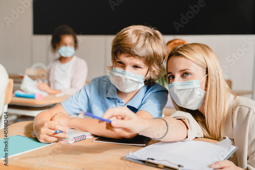 Teacher helping her student pupil schoolboy explaining material new topic task module test wearing protective face mask against Covid19 coronavirus. School education during pandemic.
