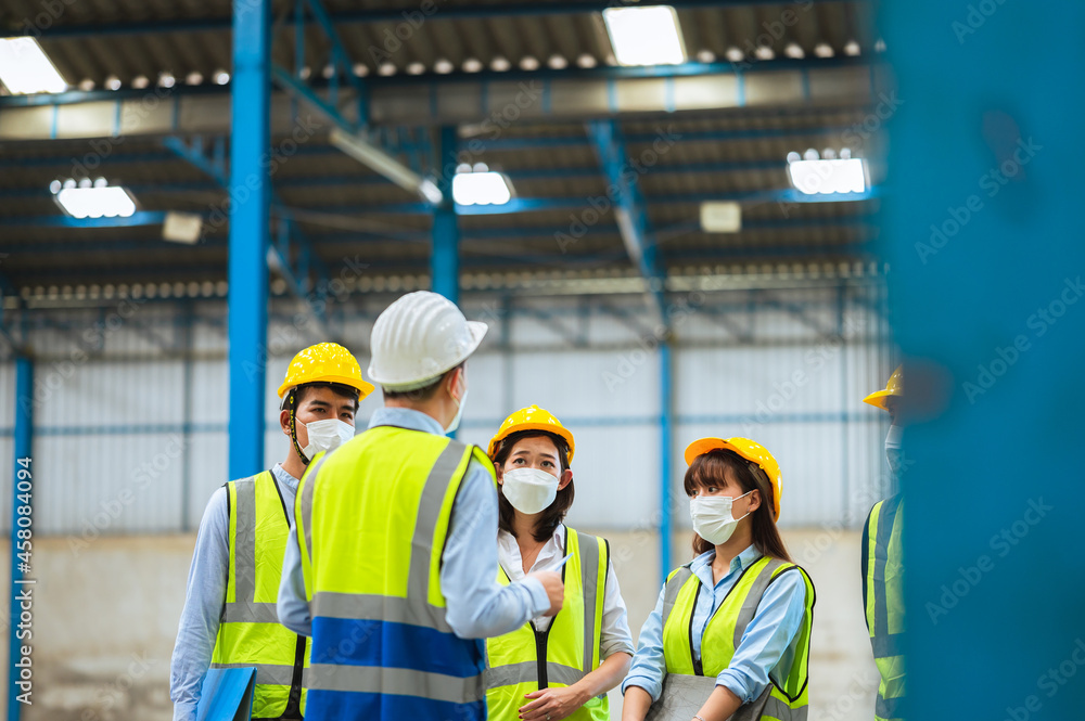The factory employee consists of engineers, foreman, technicians, and related department staff. Wear a mask, hard hat, and vest. meeting before starting work inside the warehouse. Teamwork concept. The factory employee consists of engineers, foreman, technicians, and related department staff. Wear a mask, hard hat, and vest. meeting before starting work inside the warehouse. Teamwork concept.