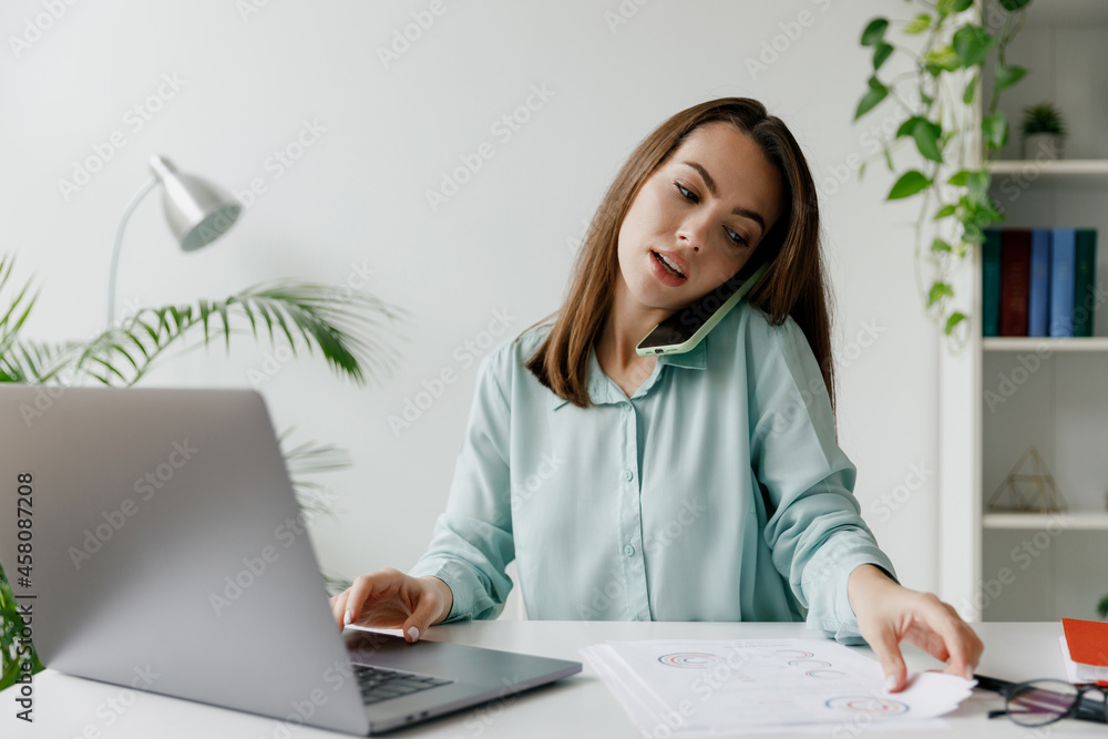 Young successful employee business woman in blue shirt talk on mobile cell phone sit work at workplace white desk with laptop pc computer take documents at office indoors. Achievement career concept. Young successful employee business woman in blue shirt talk on mobile cell phone sit work at workplace white desk with laptop pc computer take documents at office indoors. Achievement career concept.