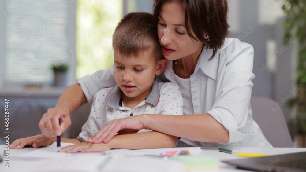 Lovely mother drawing in album with little son while sitting together at desk. Happy caucasian family using colorful pencils during creative process at home. Lovely mother drawing in album with little son while sitting together at desk. Happy caucasian family using colorful pencils during creative process at home.