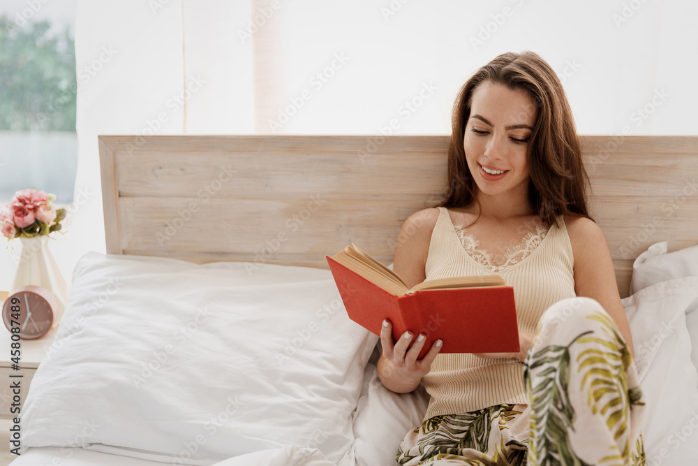 Smiling charming young woman in casual casual clothes lay on bed holding reading book rest relax spend time in bedroom lounge home in own room house be lost in reverie good day. Real estate concept. Smiling charming young woman in casual casual clothes lay on bed holding reading book rest relax spend time in bedroom lounge home in own room house be lost in reverie good day. Real estate concept.
