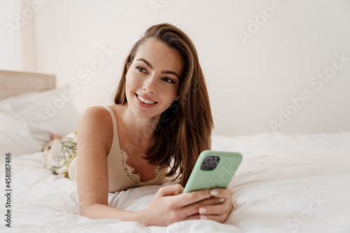 Happy young woman in casual casual clothes lying on her stomach hold in hand use mobile cell phone look aside rest relax spend time in bedroom lounge home own room house be lost in reverie good day