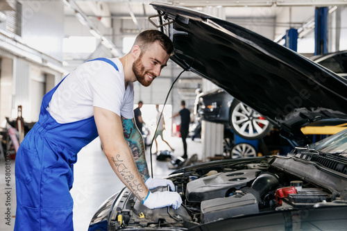 Young male professional technician car mechanic man in denim blue overalls white t-shirt gloves fixing problem with raised hood work in modern vehicle repair shop workshop indoor Tattoo translate fun.