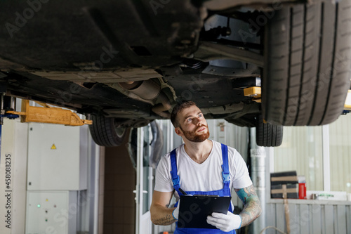 Young technician mechanic man wears denim overalls use hold clipboard papers document writing estimate outlay stand near car lift check technical condition work in vehicle repair shop workshop indoor.
