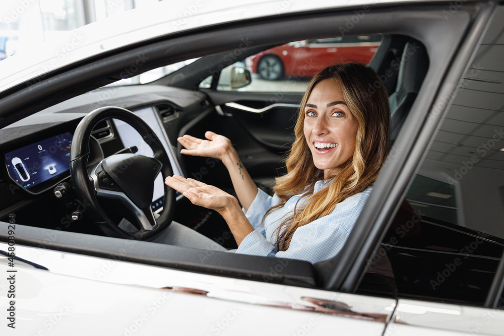 Happy woman customer buyer client in blue shirt drive without hold steering wheel show autopilot choose auto want buy new automobile in car showroom vehicle salon dealership store motor show indoor.