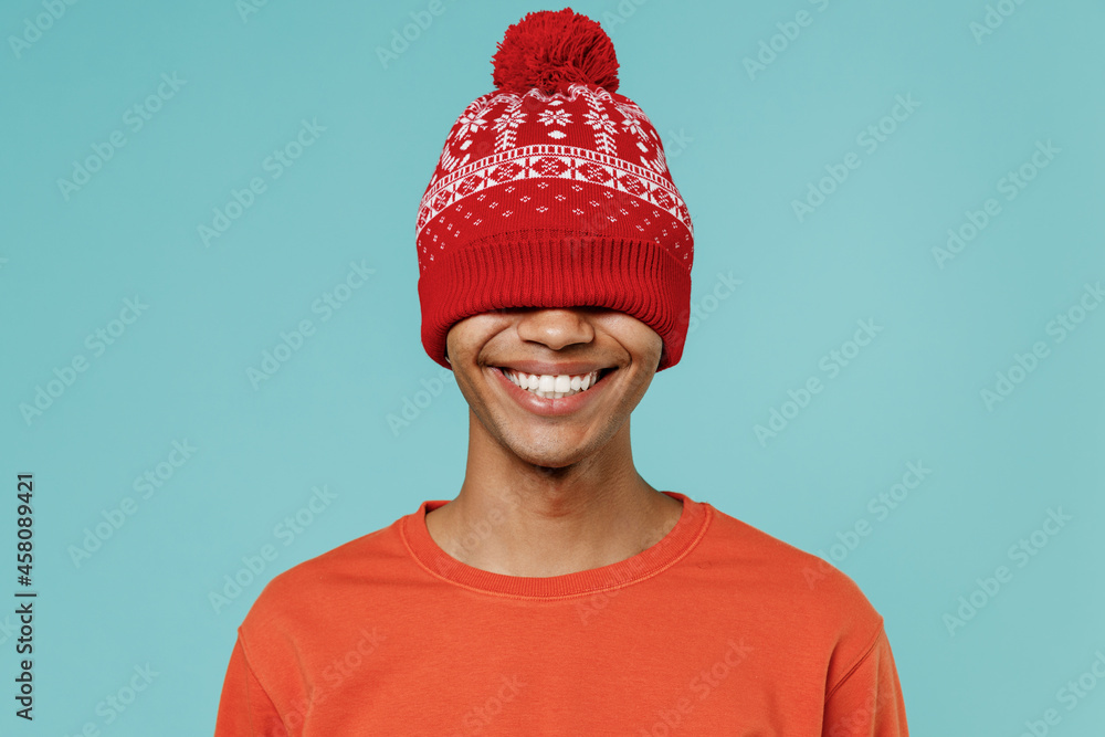 Young excited happy african american man 20s in red shirt New Year hat smiling laughing covering eyes isolated on plain pastel light blue background studio portrait. People winter lifestyle concept. Young excited happy african american man 20s in red shirt New Year hat smiling laughing covering eyes isolated on plain pastel light blue background studio portrait. People winter lifestyle concept.