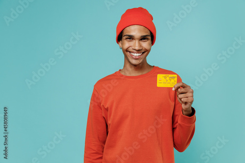 Young smiling cheerful happy cool african american man 20s in orange shirt hat hold in hand credit bank card isolated on plain pastel light blue background studio portrait. People lifestyle concept. © ViDi Studio