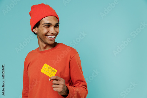 Young smiling happy cool african american man 20s in orange shirt hat hold in hand credit bank card look aside isolated on plain pastel light blue background studio portrait. People lifestyle concept. © ViDi Studio