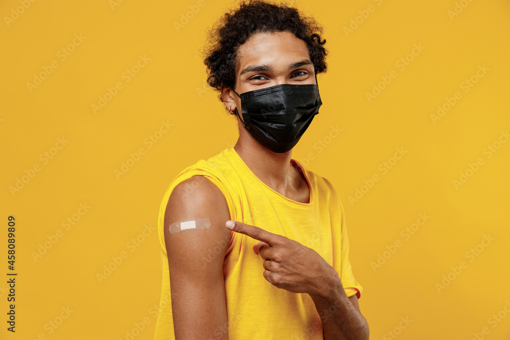Young smiling happy satisfied african american man in mask point on adhesive plaster after covid-19 coronavirus vaccine injection isolated on plain yellow background studio. People healthcare concept. Young smiling happy satisfied african american man in mask point on adhesive plaster after covid-19 coronavirus vaccine injection isolated on plain yellow background studio. People healthcare concept.
