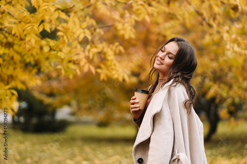 A beautiful pretty young woman in a coat drinks hot coffee walks in the autumn park smiles communicates online and talks using a mobile phone in nature in the fall forest, selective focus