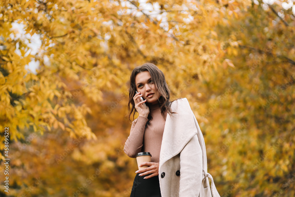 A happy woman in casual clothes smiles talks walks drinks coffee communicates chat online takes a selfie using a mobile phone technology alone in the autumn forest in nature in fall, selective focus A happy woman in casual clothes smiles talks walks drinks coffee communicates chat online takes a selfie using a mobile phone technology alone in the autumn forest in nature in fall, selective focus