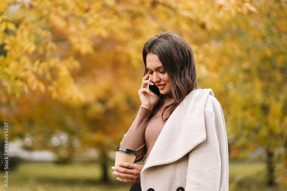 A happy woman in casual clothes smiles talks walks drinks coffee communicates chat online takes a selfie using a mobile phone technology alone in the autumn forest in nature in fall, selective focus A happy woman in casual clothes smiles talks walks drinks coffee communicates chat online takes a selfie using a mobile phone technology alone in the autumn forest in nature in fall, selective focus