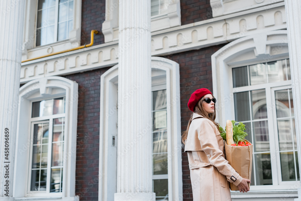 pretty young woman in trench coat, sunglasses and red beret holding paper bag with groceries near building