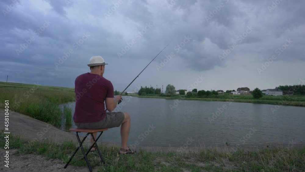 adult man fishing sitting on chair on shore of pond in summer cloudy day view from behind adult man fishing sitting on chair on shore of pond in summer cloudy day view from behind