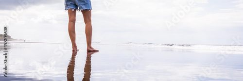 Legs of a woman standing on a beach with her silhouette reflected in the water.