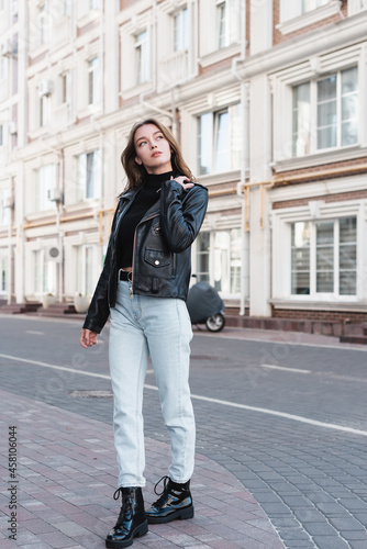 full length of stylish young woman in black turtleneck and leather jacket on urban street of europe