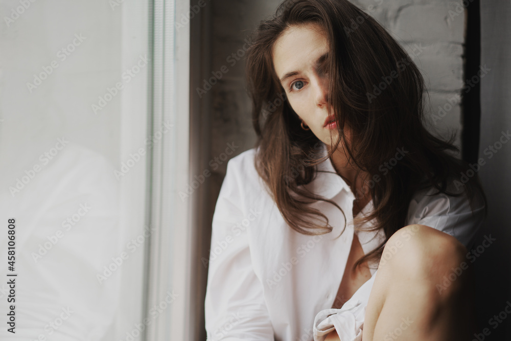 Portrait of a young beautiful sexy woman in a white shirt sitting by the window in an apartment Portrait of a young beautiful sexy woman in a white shirt sitting by the window in an apartment