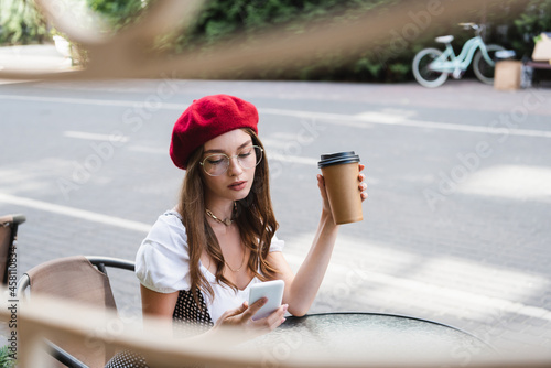 young woman in red beret and eyeglasses holding paper cup and using smartphone on terrace of cafe